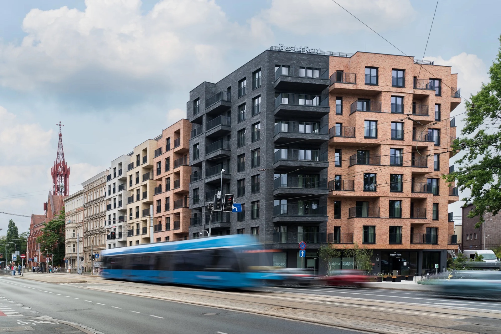 Nowy budynek mieszkalny przy ulicy, przejeżdża tramwaj, w tle kościół.